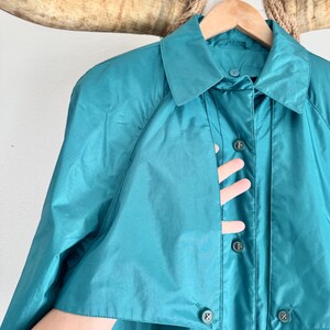 May include: Teal-colored vintage raincoat with a collared neckline and button-down closure. The coat is hanging on a wooden hanger. The coat's fabric has a slight sheen, and the buttons are a darker shade of teal.