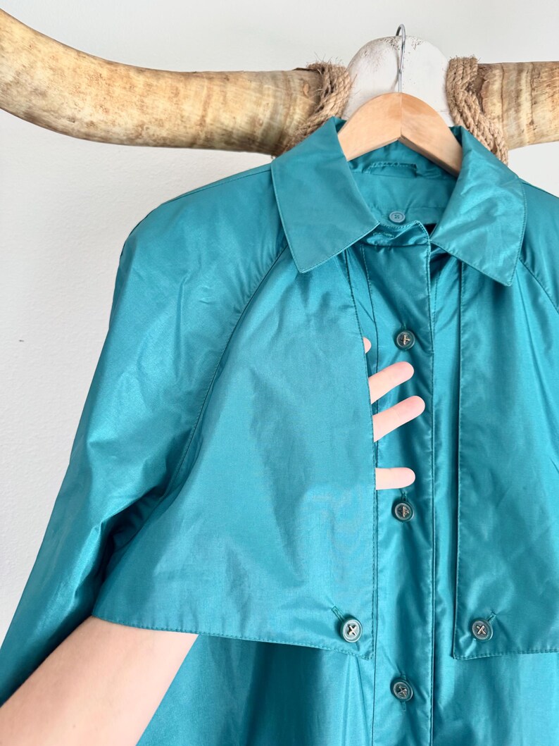 May include: Teal-colored vintage raincoat with a collared neckline and button-down closure. The coat is hanging on a wooden hanger. The coat's fabric has a slight sheen, and the buttons are a darker shade of teal.