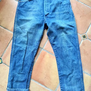 May include: Blue denim jeans with a classic five-pocket design. The jeans have a straight leg cut and a button and zipper closure. The cuffs are rolled up, revealing the lighter-colored interior fabric. The jeans are made of a durable denim material.