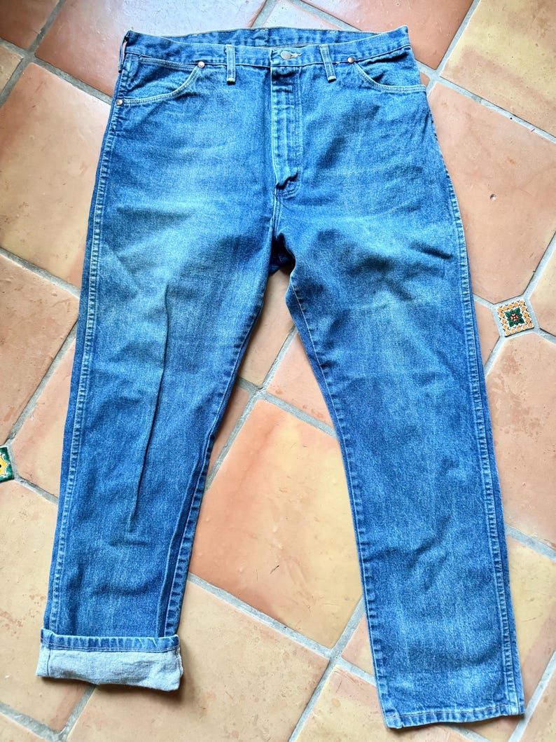 May include: Blue denim jeans with a classic five-pocket design. The jeans have a straight leg cut and a button and zipper closure. The cuffs are rolled up, revealing the lighter-colored interior fabric. The jeans are made of a durable denim material.