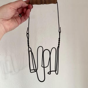 May include: A vintage black wire bottle carrier with a wooden handle. The carrier has a looped design to hold bottles and is suspended by thin wire. The wooden handle is a light brown color.