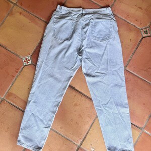 May include: Light blue denim jeans, viewed from the back, with two back pockets and a waistband. The jeans are laid flat on a terracotta tile surface. The denim appears to be a light wash.
