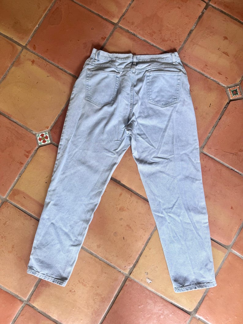 May include: Light blue denim jeans, viewed from the back, with two back pockets and a waistband. The jeans are laid flat on a terracotta tile surface. The denim appears to be a light wash.
