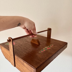 May include: A vintage wooden sewing box with a handle and a hinged lid. The box is a rich brown color with floral accents in red, green, and yellow. The box is being held by a person.