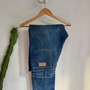 May include: Blue denim jeans hanging on a wooden hanger. The jeans feature a classic five-pocket design, a brown leather patch, and the Wrangler logo. The jeans are displayed against a white wall with a cactus and a skull.