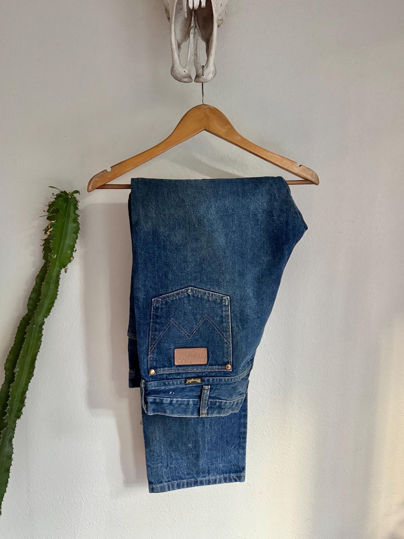 May include: Blue denim jeans hanging on a wooden hanger. The jeans feature a classic five-pocket design, a brown leather patch, and the Wrangler logo. The jeans are displayed against a white wall with a cactus and a skull.