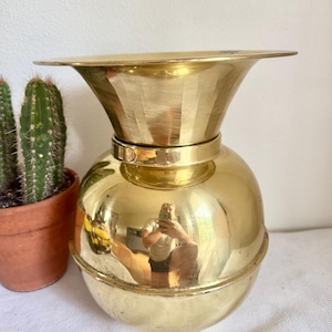 May include: A shiny, gold-colored metal vase with a wide, flared top and a rounded body. The vase has a reflective surface, and is sitting on a white surface. A potted cactus is visible in the background.