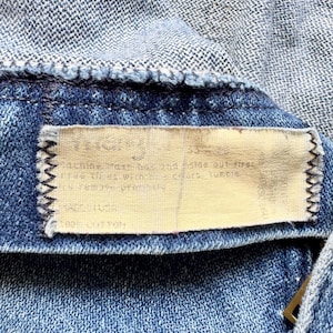 K&ouml;nnte beinhalten: Nahaufnahme eines Jeanskleidungsst&uuml;cks, das die Textur und die N&auml;hte zeigt. Ein rechteckiges Etikett mit Text ist in das Kleidungsst&uuml;ck eingen&auml;ht. Der Denim ist mittelblau, mit einem helleren blauen Stoff oben sichtbar.
