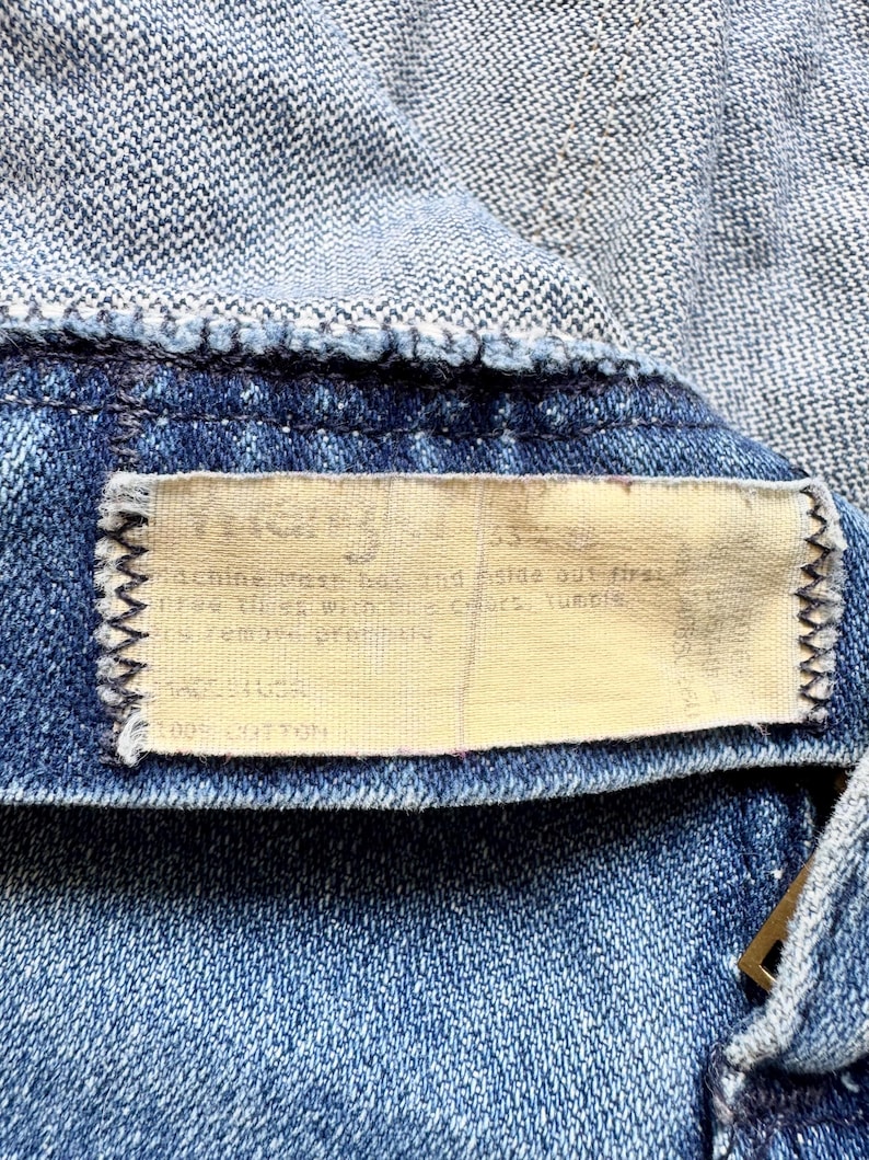 K&ouml;nnte beinhalten: Nahaufnahme eines Jeanskleidungsst&uuml;cks, das die Textur und die N&auml;hte zeigt. Ein rechteckiges Etikett mit Text ist in das Kleidungsst&uuml;ck eingen&auml;ht. Der Denim ist mittelblau, mit einem helleren blauen Stoff oben sichtbar.