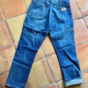 May include: Blue denim jeans with a classic five-pocket design. The jeans have a faded wash and a leather patch on the back pocket. The cuffs are rolled up, and the jeans are laid flat on a tiled surface.