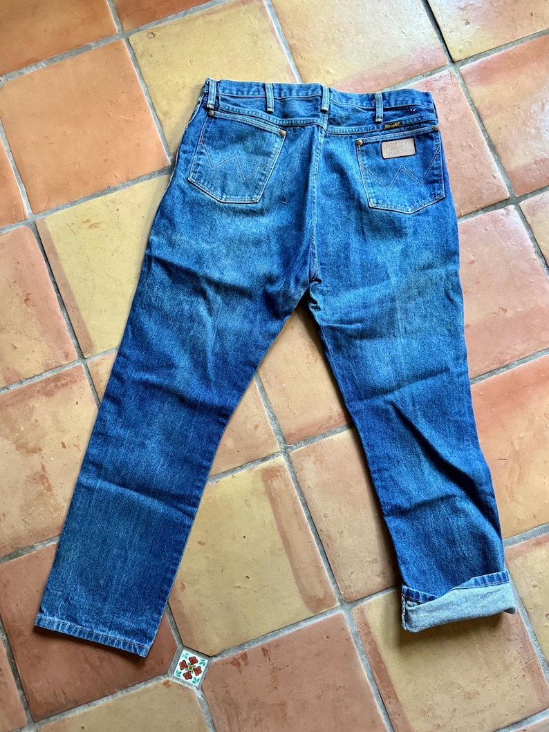 May include: Blue denim jeans with a classic five-pocket design. The jeans have a faded wash and a leather patch on the back pocket. The cuffs are rolled up, and the jeans are laid flat on a tiled surface.