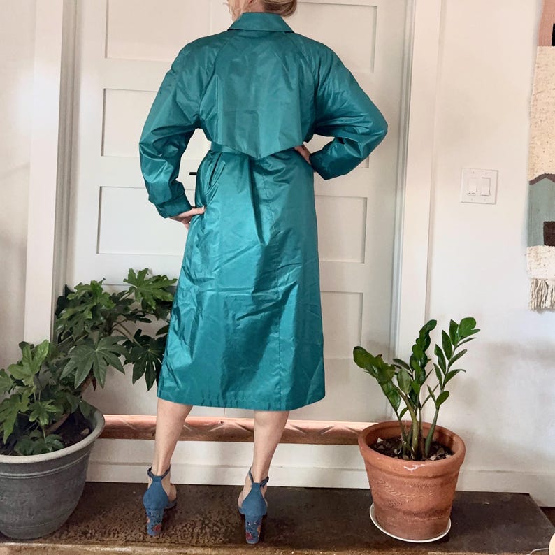 May include: Teal-colored vintage raincoat with a collared neckline, long sleeves, and a belted waist. The knee-length coat is made of a shiny, water-resistant material. The person is wearing blue heels.