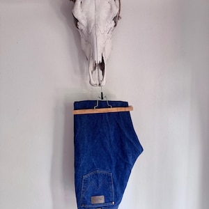 May include: A pair of dark blue denim jeans featuring a classic five-pocket design, a leather-like patch on the back waistband, and signature stitching on the rear pocket, suspended from a wooden hanger against a plain white wall.