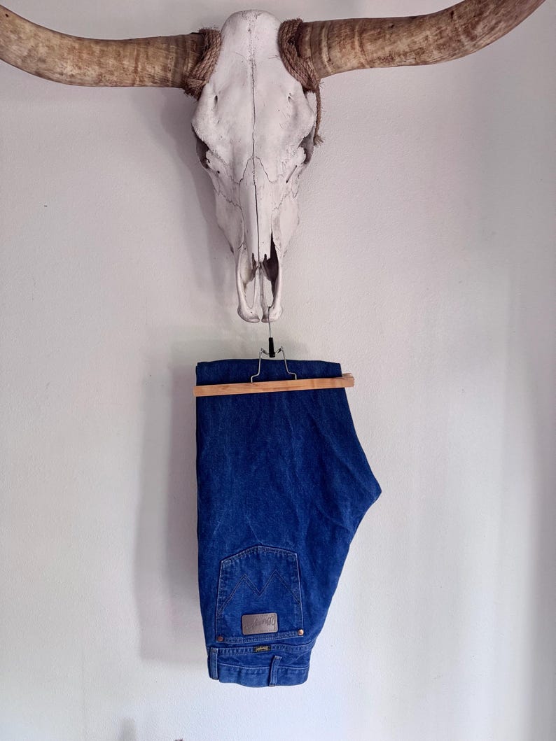 May include: A pair of dark blue denim jeans featuring a classic five-pocket design, a leather-like patch on the back waistband, and signature stitching on the rear pocket, suspended from a wooden hanger against a plain white wall.