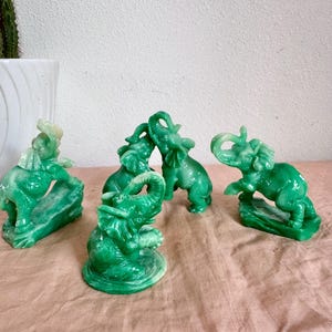 May include: A collection of four small, translucent green elephant figurines. Each elephant is depicted in a different pose, with some standing on rocky bases and others on circular platforms, all featuring smooth, glossy surfaces and raised trunks.