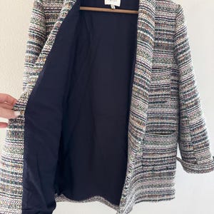 May include: A multi-colored tweed blazer with a navy blue lining. The blazer is hanging on a wooden hanger. The tweed fabric features a mix of colors, including white, green, and purple. The blazer has a classic lapel and two pockets.