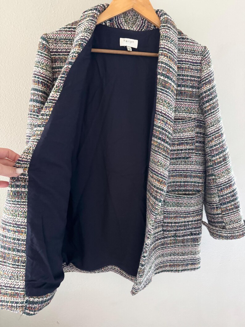 May include: A multi-colored tweed blazer with a navy blue lining. The blazer is hanging on a wooden hanger. The tweed fabric features a mix of colors, including white, green, and purple. The blazer has a classic lapel and two pockets.