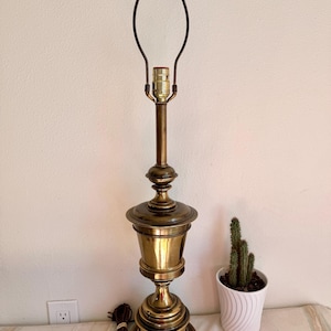May include: A vintage brass table lamp with a classic design. The lamp features a detailed base, a cylindrical body, and a slender neck leading to the bulb socket. The lamp is approximately 28 inches tall.