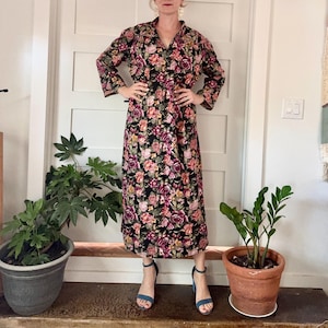 May include: A long-sleeved floral dress in a dark color palette, featuring a V-neck and a full-length skirt. The dress is adorned with a pattern of pink, peach, and purple flowers. The model is wearing blue sandals. The dress is suitable for a vintage or bohemian style.