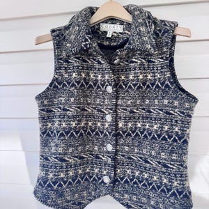 May include: A sleeveless vest with a geometric pattern in navy blue and cream. The vest has a collared neckline, decorative buttons, and is hanging on a wooden hanger. The tag reads "COUNTY CLOTHING COMPANY".