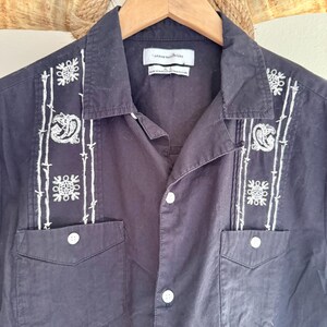 May include: Black button-down shirt with white embroidered detailing. The shirt features two chest pockets and a collared neckline. The embroidery includes floral and paisley designs. The tag reads "Urban Outfitters".