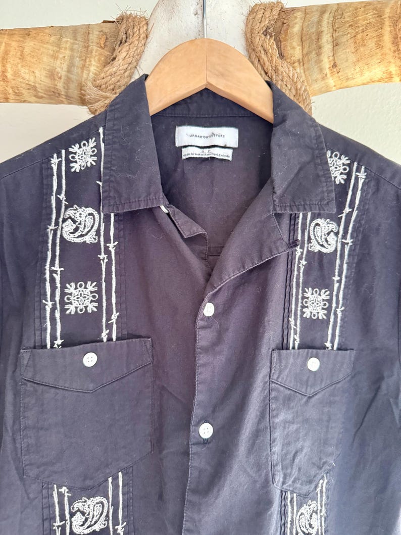 May include: Black button-down shirt with white embroidered detailing. The shirt features two chest pockets and a collared neckline. The embroidery includes floral and paisley designs. The tag reads "Urban Outfitters".
