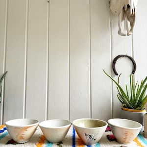 May include: Four small ceramic bowls with different designs. The bowls are white with various patterns in shades of brown, orange, and green. The bowls are arranged in a row on a colorful striped rug.