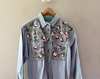 May include: Light blue button-down shirt with long sleeves, featuring embroidered Texas flags and floral designs on the yoke and pockets. The shirt is hanging on a wooden hanger.