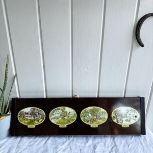 May include: A black metal wall decor with four oval-shaped illustrations depicting the four seasons. The illustrations are in a vintage style and feature a farmhouse with trees and other details. The seasons are labeled "Spring", "Summer", "Autumn", and "Winter".