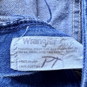 May include: A blue denim fabric garment featuring a white rectangular interior tag with the Wrangler brand name printed in black. The tag includes care instructions, the text Made in USA, and 100% Cotton, with a handwritten PT mark.