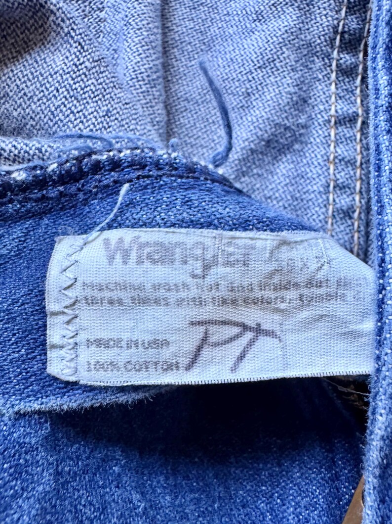 May include: A blue denim fabric garment featuring a white rectangular interior tag with the Wrangler brand name printed in black. The tag includes care instructions, the text Made in USA, and 100% Cotton, with a handwritten PT mark.