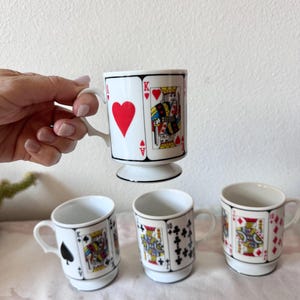 May include: Set of four white ceramic mugs featuring playing card designs. One mug is held up, displaying the King of Hearts. The other three mugs show the King of Spades, the 10 of Clubs, and the King of Diamonds.