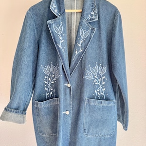 May include: A medium-wash blue denim blazer featuring white embroidered leaf and vine patterns on the lapels and front panels. The jacket includes two large patch pockets, a notched collar, and two silver-toned buttons down the center front.
