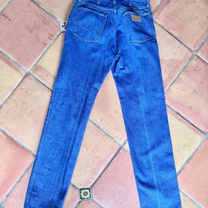 May include: Blue denim jeans with a classic five-pocket design. The jeans have a straight leg cut and a brown leather patch on the back. The jeans are laid flat on a tiled surface.
