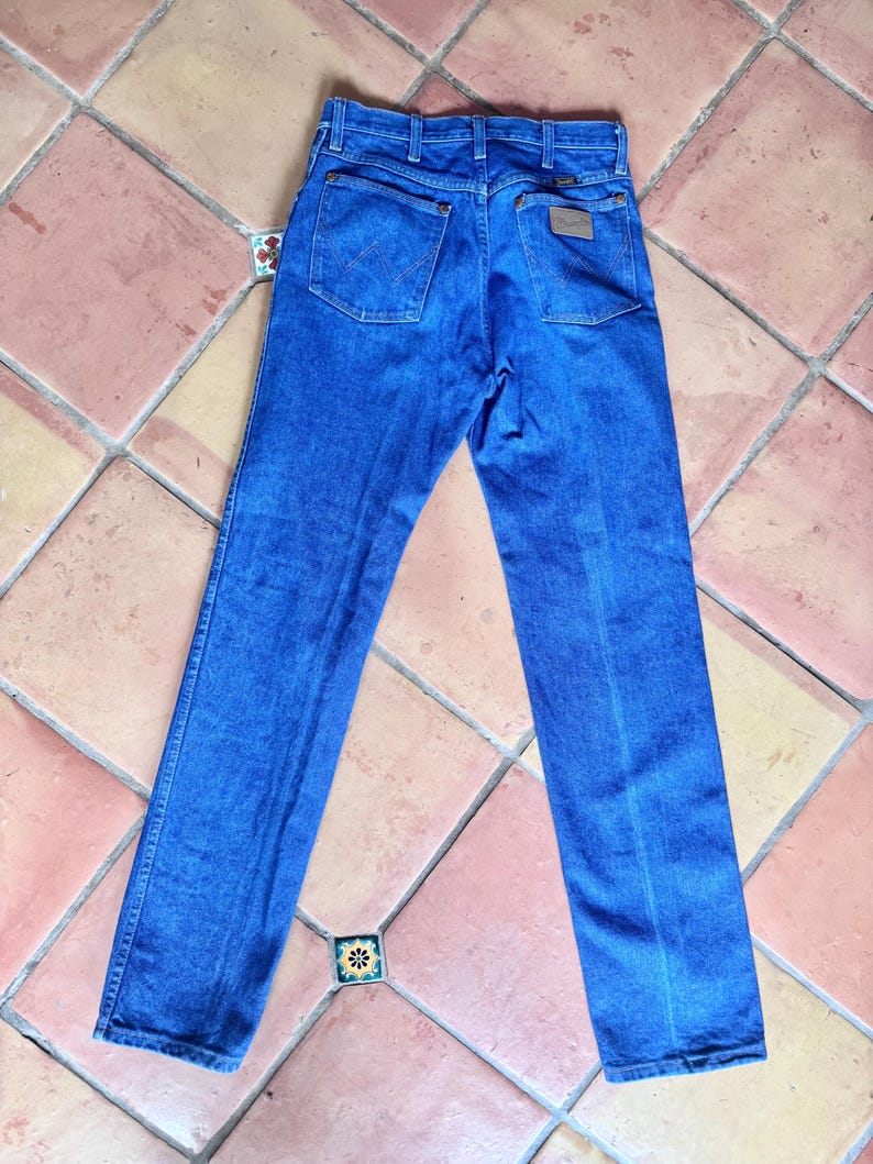 May include: Blue denim jeans with a classic five-pocket design. The jeans have a straight leg cut and a brown leather patch on the back. The jeans are laid flat on a tiled surface.