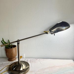 May include: A vintage-style desk lamp with a bronze finish. The lamp features an adjustable arm and a teardrop-shaped shade. The base is oval, and the lamp is plugged into a wall outlet. A small potted plant is visible in the background.