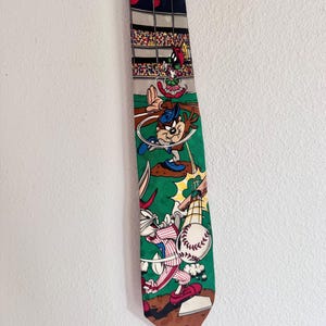 May include: A necktie featuring a colorful Looney Tunes baseball scene. The design includes Marvin the Martian in the stands, the Tasmanian Devil pitching on a mound, and Bugs Bunny batting at home plate with a baseball and bat in motion.