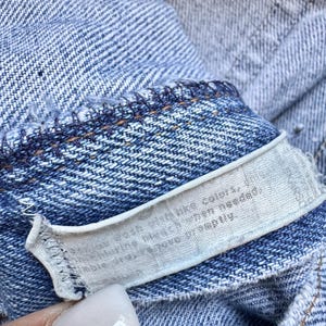 May include: Close-up of a light blue denim garment with a white tag. The tag has text that is partially visible. The denim has visible stitching and a textured weave.