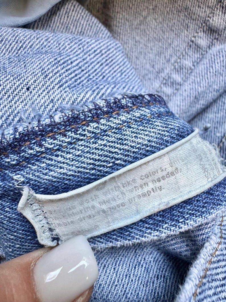 May include: Close-up of a light blue denim garment with a white tag. The tag has text that is partially visible. The denim has visible stitching and a textured weave.