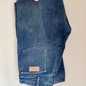 May include: Folded blue denim jeans with a classic five-pocket design. Features a brown leather patch with the "Wrangler" logo on the back pocket. The jeans have a straight leg cut and are held up by a hand.