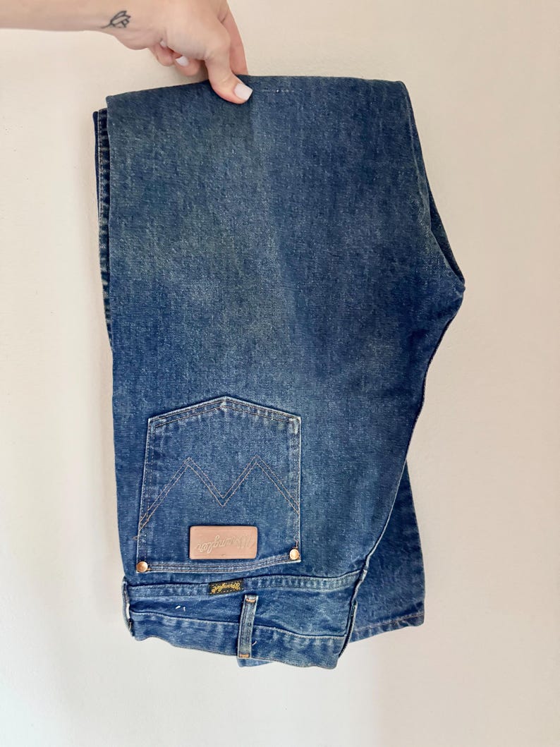 May include: Folded blue denim jeans with a classic five-pocket design. Features a brown leather patch with the "Wrangler" logo on the back pocket. The jeans have a straight leg cut and are held up by a hand.