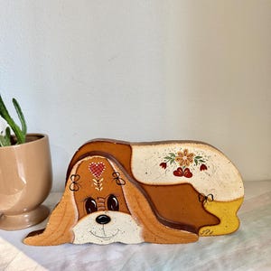 May include: A wooden dog-shaped decorative item, painted in shades of brown, cream, and yellow. The dog has large, floppy ears, a smiling face, and floral and heart accents. The item is likely a vintage or handmade piece.