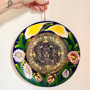 May include: A round, hand-painted decorative plate with a dark blue background. The plate features a central Aztec calendar design, surrounded by yellow calla lilies, and colorful floral accents. A red string is attached for hanging.