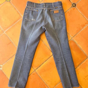 May include: Blue denim jeans with a classic five-pocket design. The jeans feature a brown leather patch on the back pocket and a metal button closure. The jeans are laid flat on an orange tile surface.
