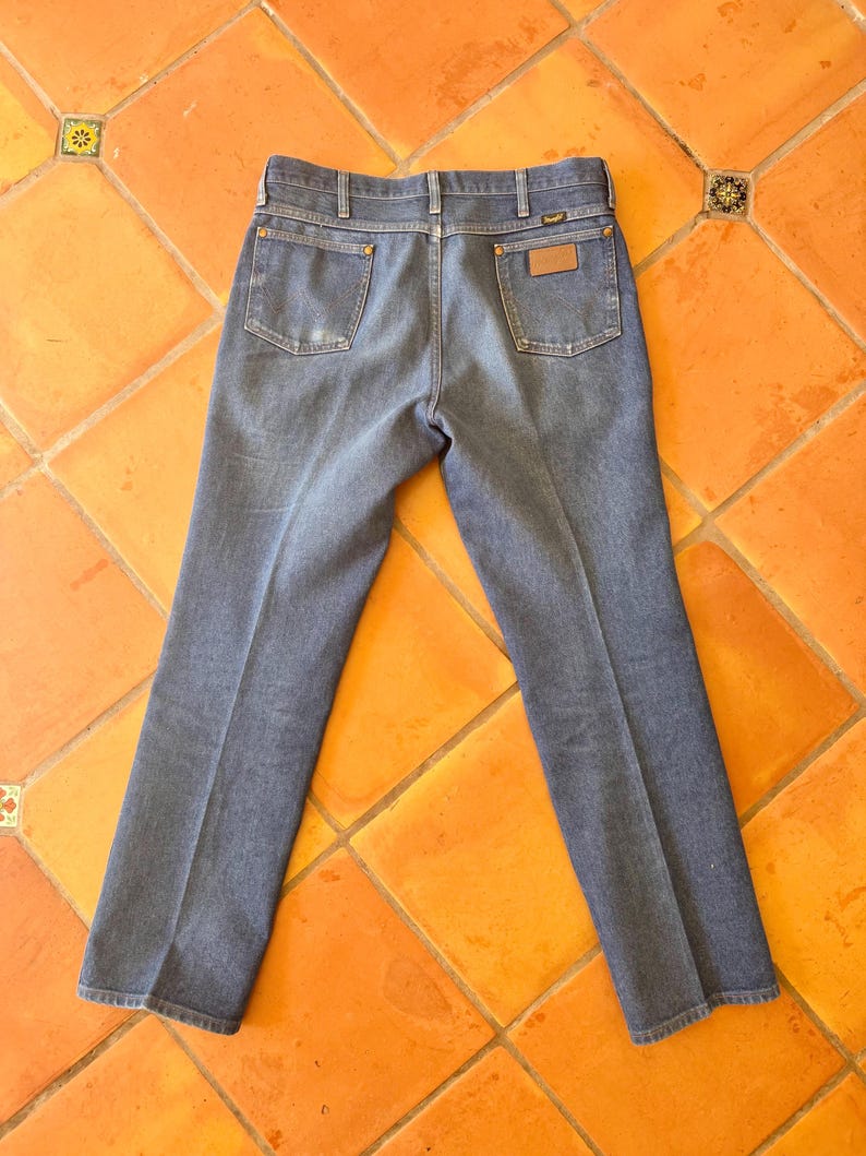 May include: Blue denim jeans with a classic five-pocket design. The jeans feature a brown leather patch on the back pocket and a metal button closure. The jeans are laid flat on an orange tile surface.