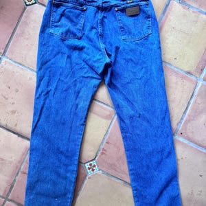 May include: A pair of blue denim jeans featuring a classic five-pocket design, a high-rise waist with belt loops, and a brown leather patch on the back right pocket. The back pockets display a signature W-shaped stitching detail in matching blue thread.