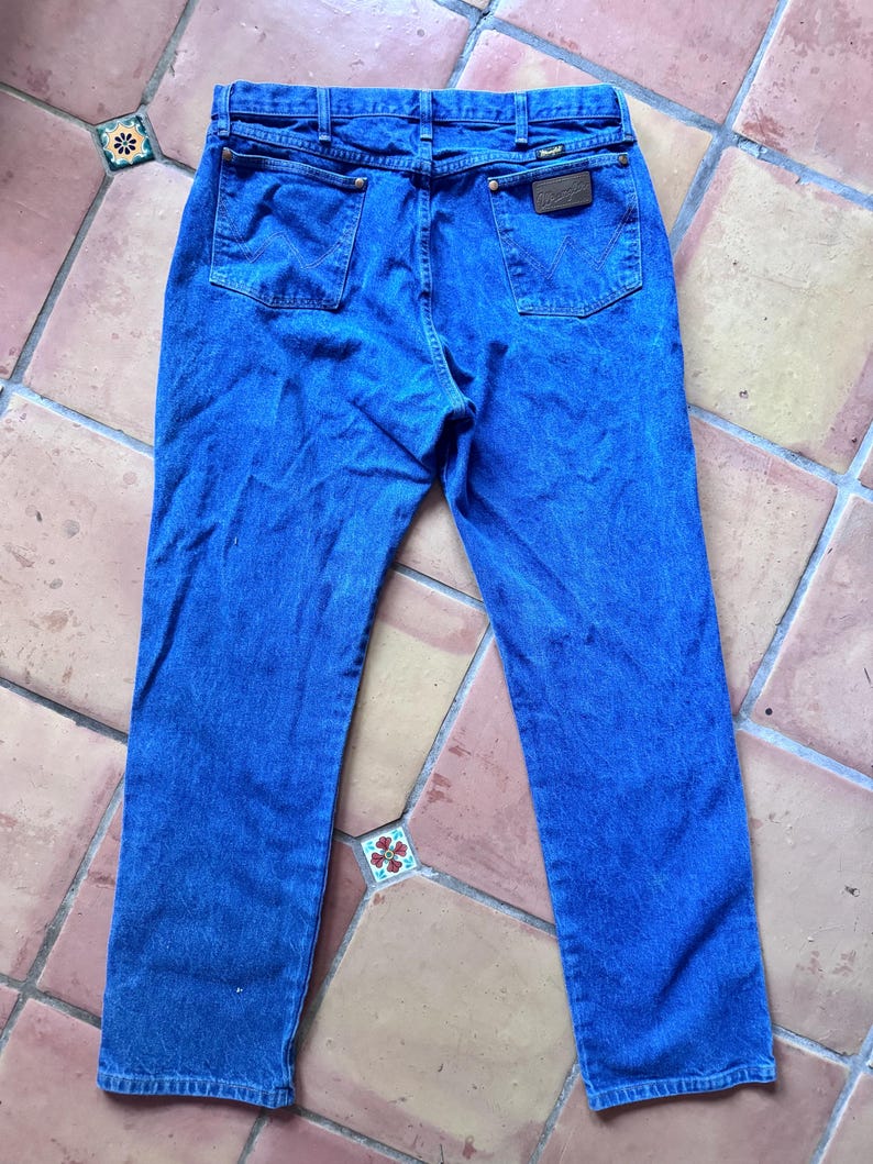 May include: A pair of blue denim jeans featuring a classic five-pocket design, a high-rise waist with belt loops, and a brown leather patch on the back right pocket. The back pockets display a signature W-shaped stitching detail in matching blue thread.