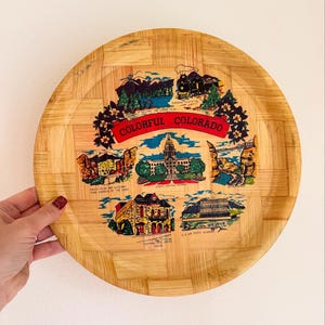 May include: A round wooden tray with a woven pattern features a colorful illustration of various landmarks in Colorado. The tray includes the text "Colorful Colorado" and names of the landmarks, including "Pikes Peak and Gateway, Three Garden of the Gods", "State Capitol, Denver", "Central City Opera House, Opera", "U.S. Air Force Academy", and "Tumbler Gorge Recreation Area".