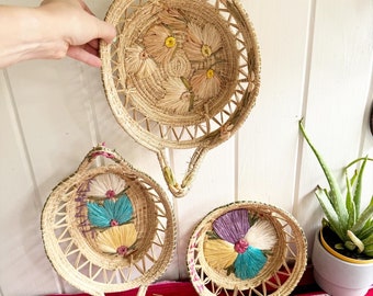 May include: Three woven straw baskets with floral designs. The largest basket has yellow flowers, the medium basket has blue, yellow, and pink flowers, and the smallest basket has purple, white, and blue flowers.