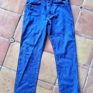 Puede incluir: Un par de vaqueros de mezclilla azul medio con un dise&ntilde;o cl&aacute;sico de cinco bolsillos, cierre de botones y remaches de color cobre en las esquinas de los bolsillos. Los vaqueros tienen un corte recto con costuras visibles en las piernas.